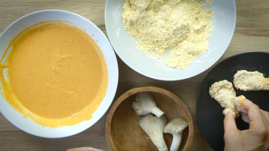 White bowl with yellow batter, white bowl with flour mix, brown bowl with oyster mushrooms. A hand is holding a breaded piece.