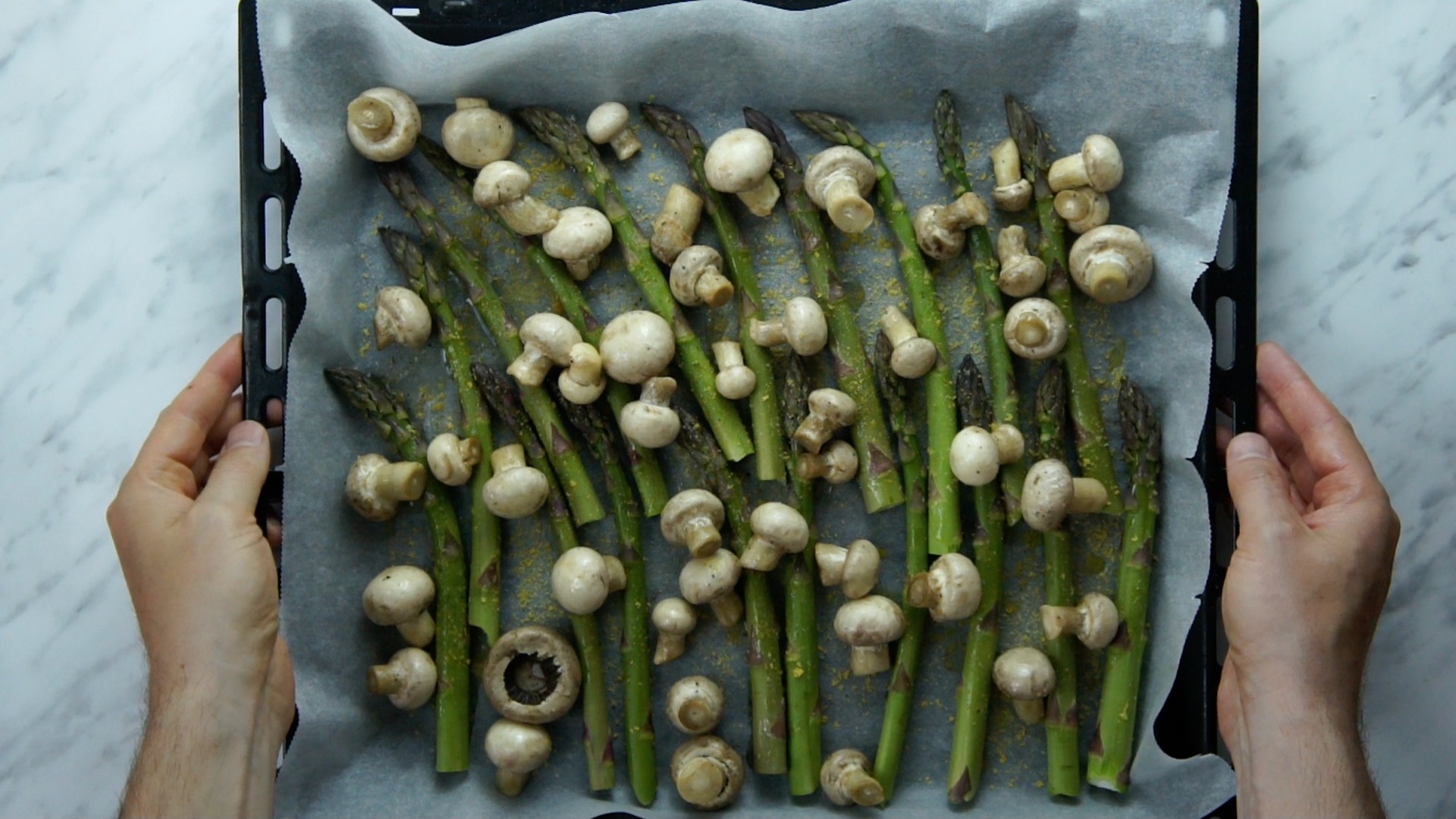 Green asparagus and small mushrooms on a baking sheet and parchment paper sprinkled with salt, black pepper, and yellow flakes.