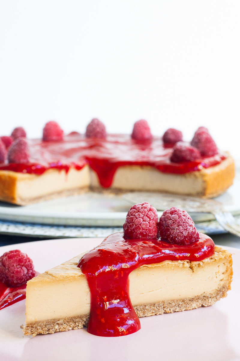 One slice of cheesecake with vibrant red sauce and raspberries on top is served on a light pink plate. The whole cake is in the background.