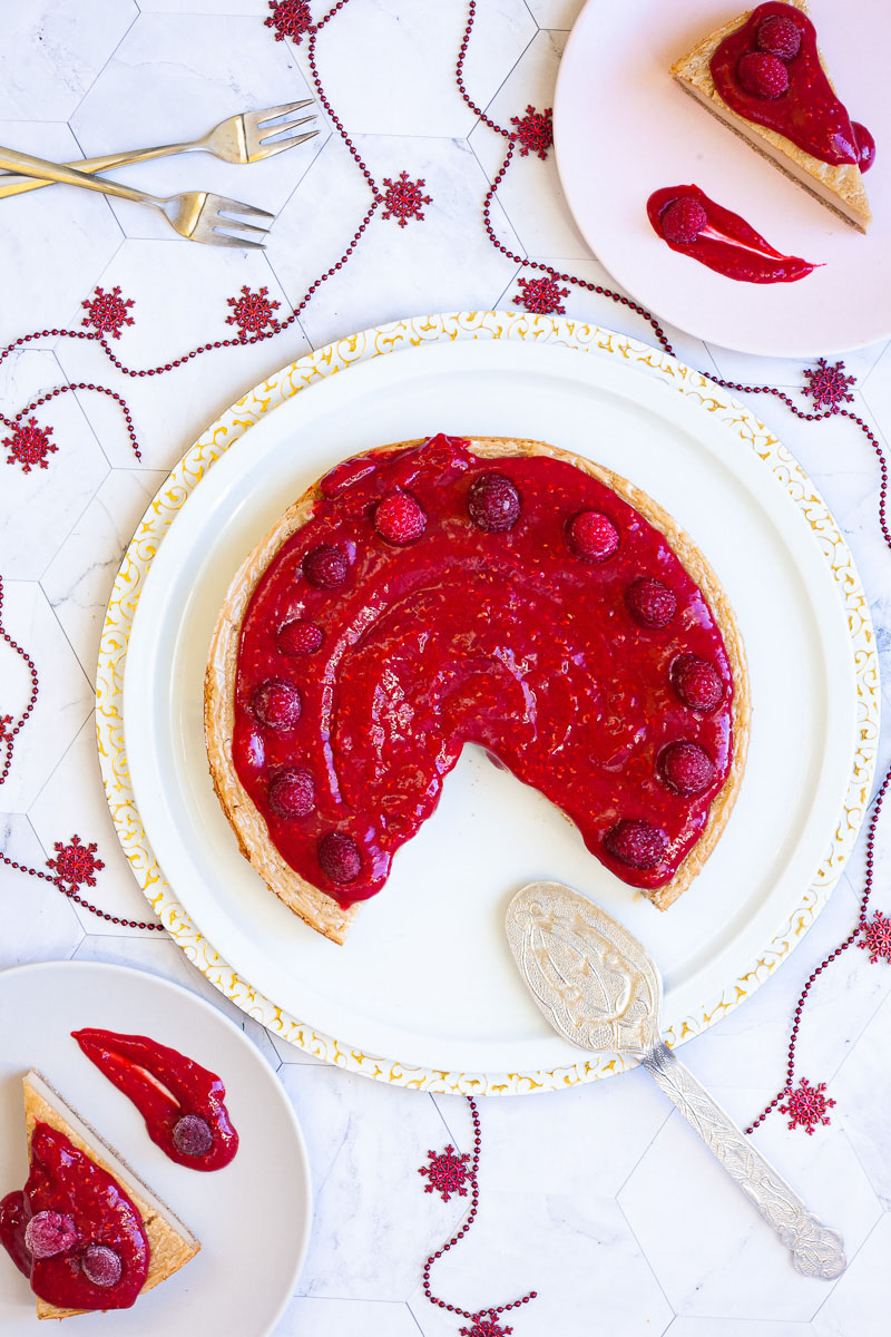 A cheesecake with vibrant red sauce and raspberries on top is pictured from above. 2 slices are missing which are served on a light pink plates next to it.