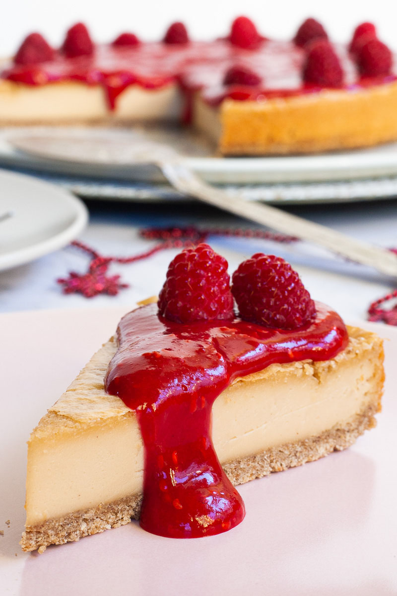 One slice of cheesecake with vibrant red sauce and raspberries on top is served on a light pink plate. The whole cake is in the background.