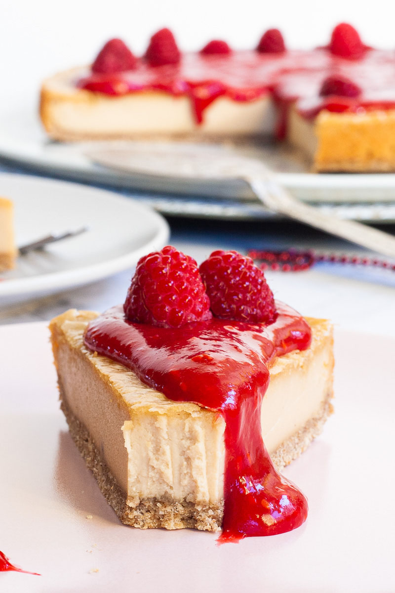 One slice of cheesecake with vibrant red sauce and raspberries on top is served on a light pink plate. The whole cake is in the background.