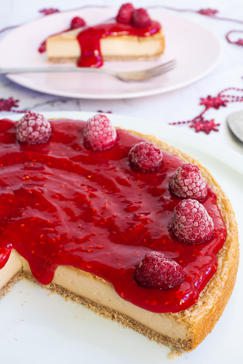 A light yellow cheesecake with vibrant red sauce and raspberries on top is placed on a large round white plate. One slice on a pink plate is behind it.