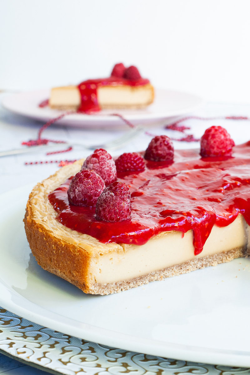 A light yellow cheesecake with vibrant red sauce and raspberries on top is placed on a large round white plate. One slice on a pink plate is behind it.