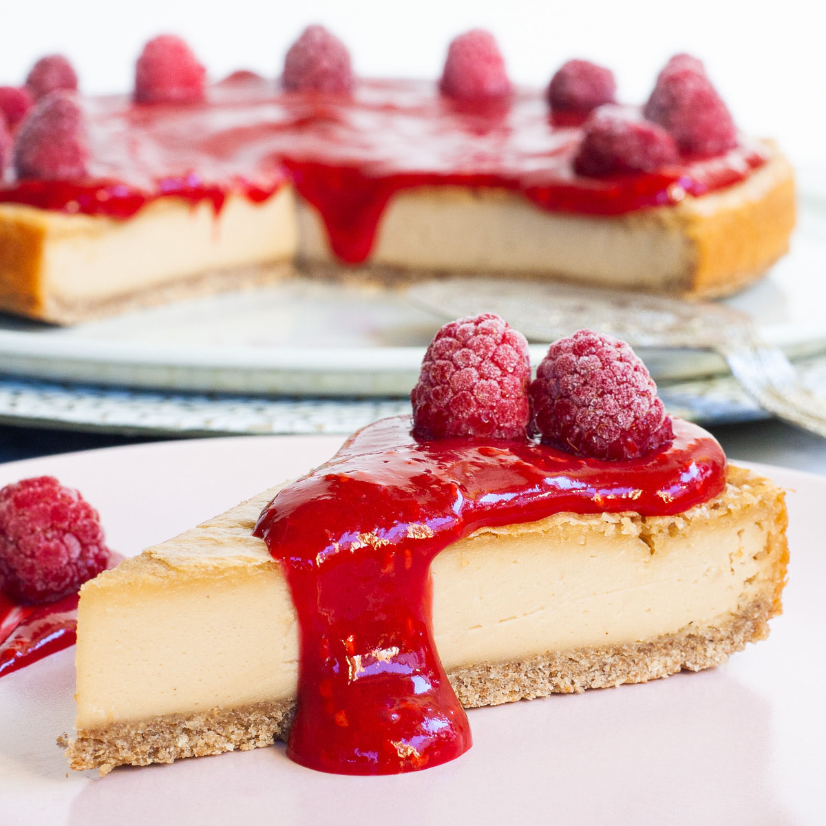 One slice of cheesecake with vibrant red sauce and raspberries on top is served on a light pink plate. The whole cake is in the background.