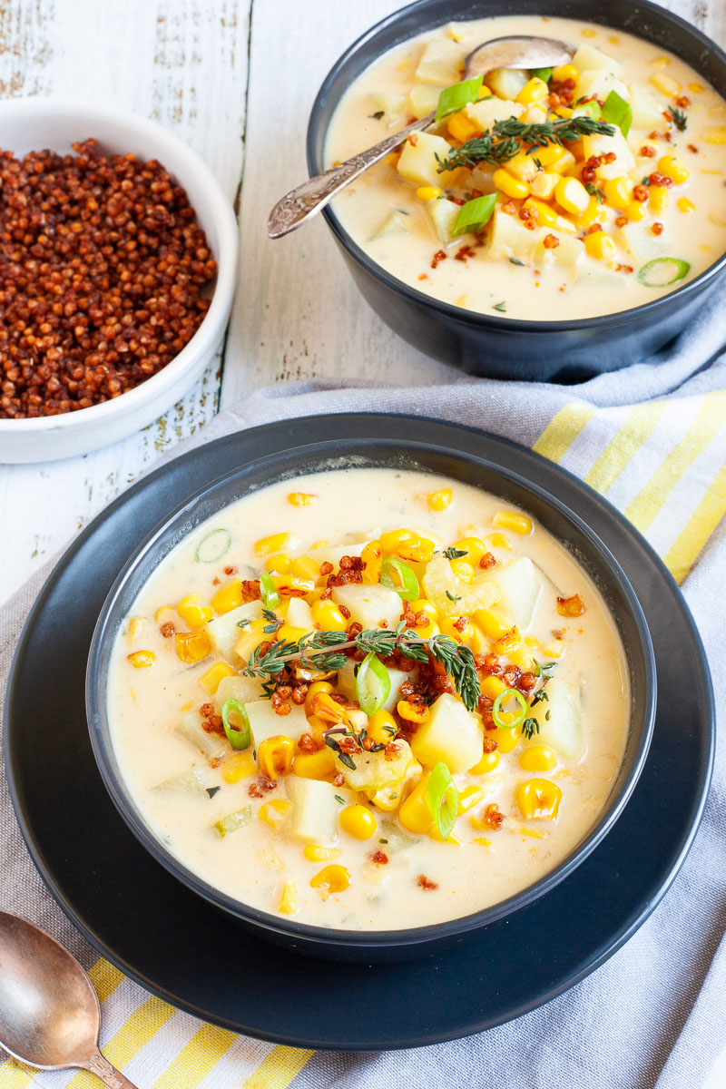 Light yellow soup served in two black bowls with croutons, corn, and diced potatoes. It is sprinkled with freshly chopped herbs, brown bacon bits and sliced spring onion. A small white bowl is full of brown bacon bits.