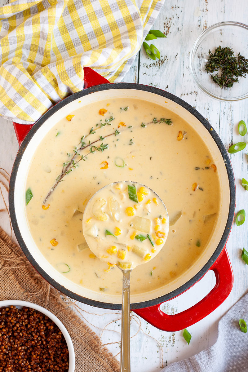 Red dutch oven from above with a light yellow soup. A laddle is taking a serving with visible diced potatoes, corn and celery slices. A small white bowl is next to it full of brown bacon bits.