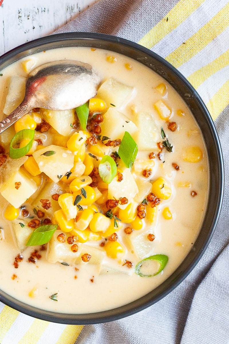 Light yellow soup served in a black bowl with croutons, corn, and diced potatoes. It is sprinkled with freshly chopped herbs, brown bacon bits and sliced spring onion.