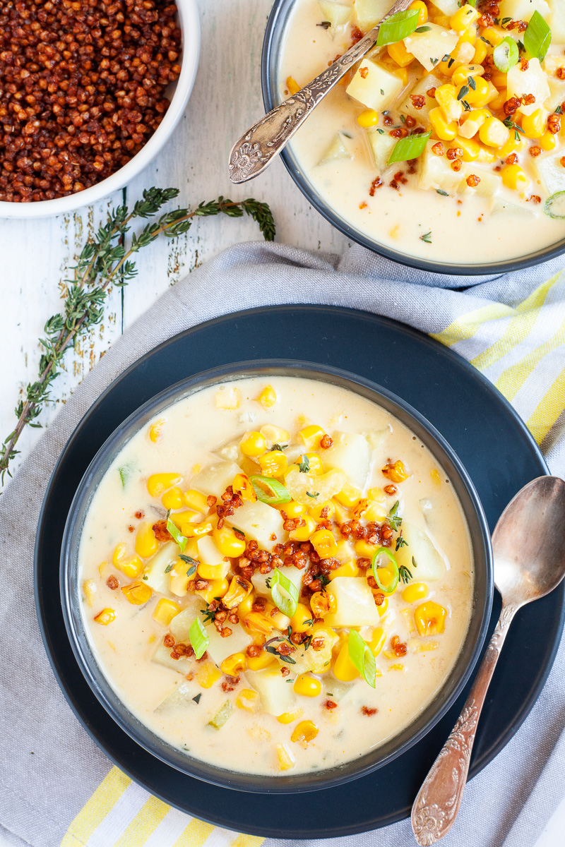 Two black bowls with light yellow soup topped with corn, spring onion rings, brown bacon bits and fresh herbs. 