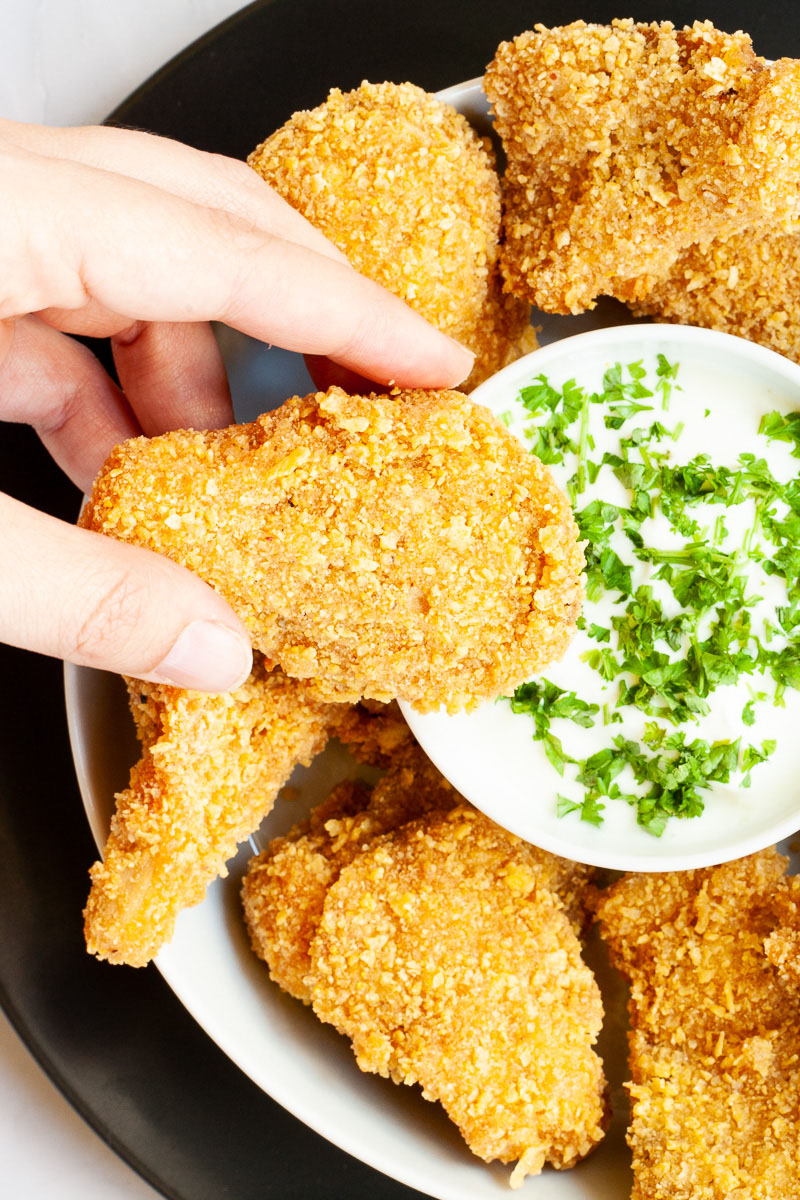Brown breaded nuggets in a round plate with a white sauce in the middle with chopped green herbs. A hand is holding a piece
