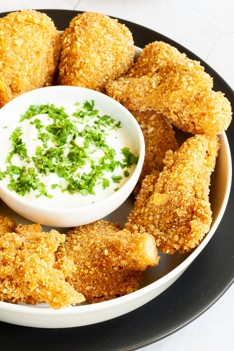 Brown breaded nuggets in a round plate with a white sauce in the middle with chopped green herbs.
