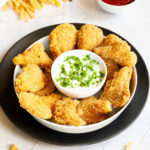 Brown breaded nuggets in a round plate with a white sauce in the middle with chopped green herbs. French fries on the side and a small bowl of ketchup