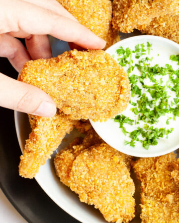 Brown breaded nuggets in a round plate with a white sauce in the middle with chopped green herbs. A hand is holding a piece