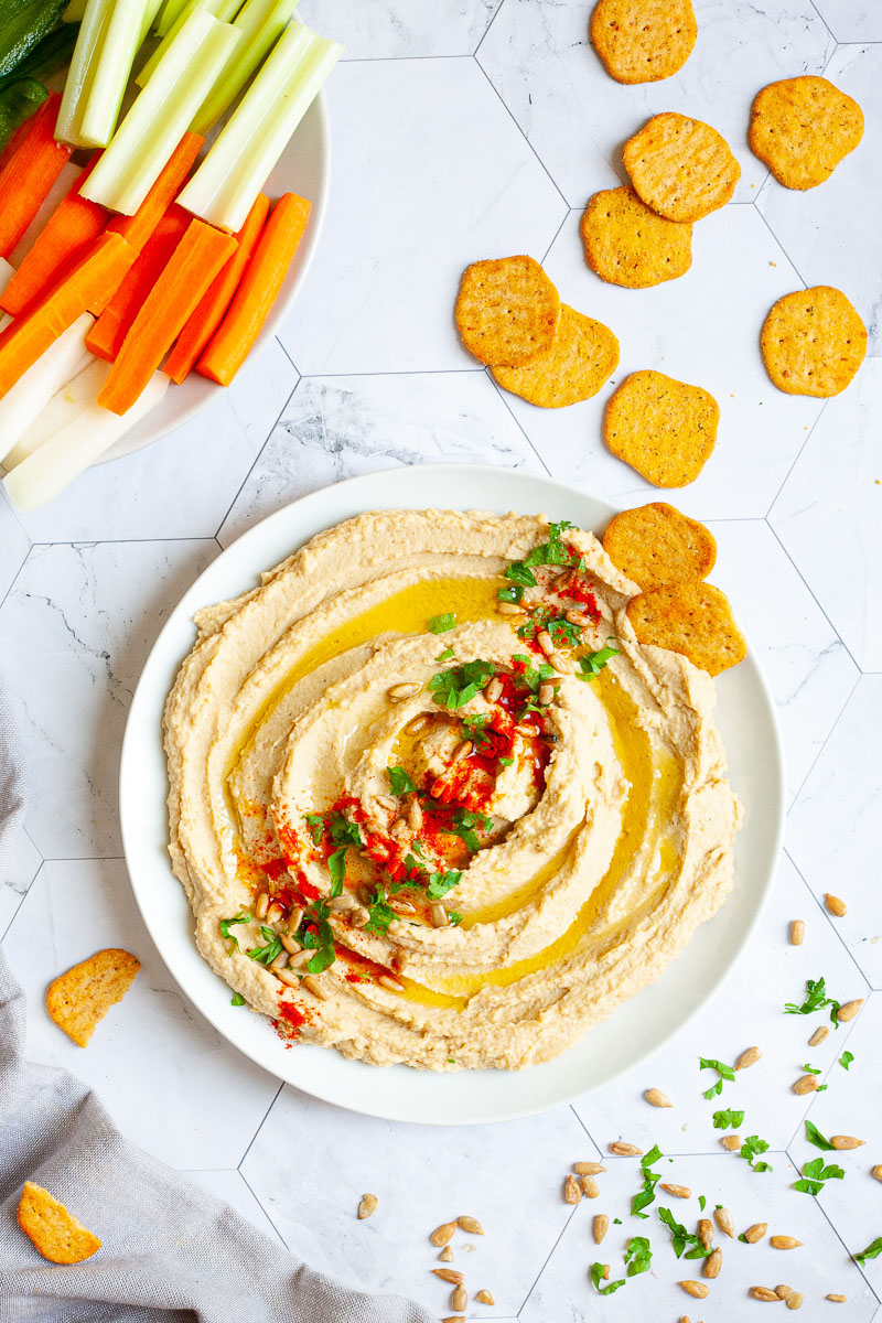 White plate with light brown hummus sprinkled on top with sunflower seeds, chopped parsley and paprika powder. Crackers are scattered around it and there is a small plate in the corner with carrot and celery sticks.