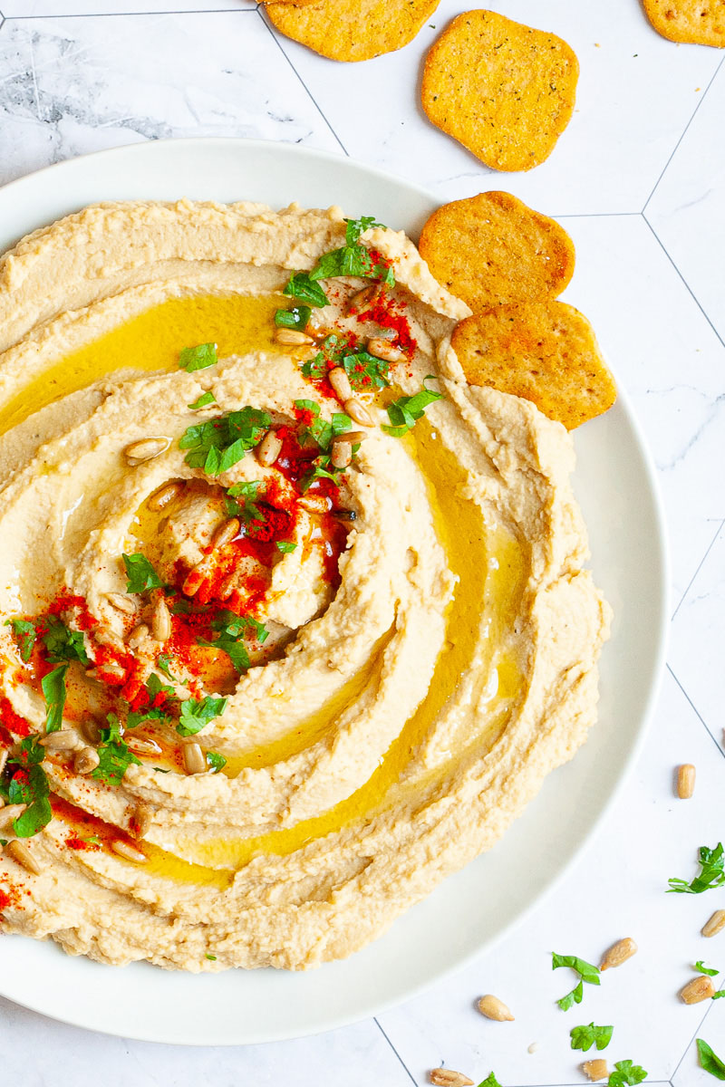 White plate with light brown hummus sprinkled on top with sunflower seeds, chopped parsley and paprika powder. Crackers are scattered around it.