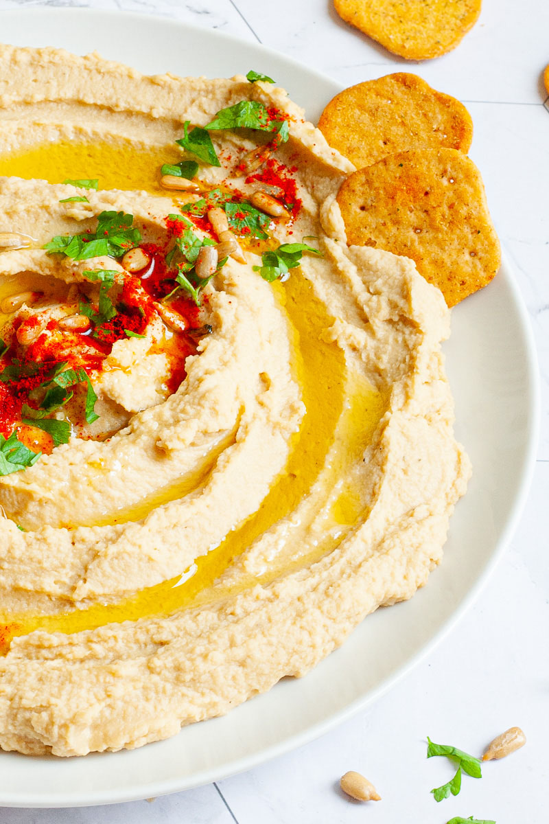 White plate with light brown hummus sprinkled on top with sunflower seeds, chopped parsley and paprika powder.