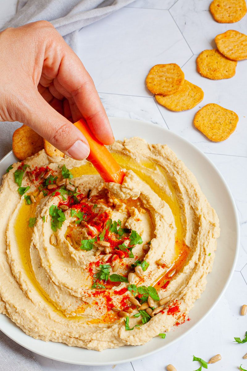 White plate with light brown hummus sprinkled on top with sunflower seeds, chopped parsley and paprika powder. Crackers are scattered around it. A hand is holding a carrot stick and dips it in the hummus.