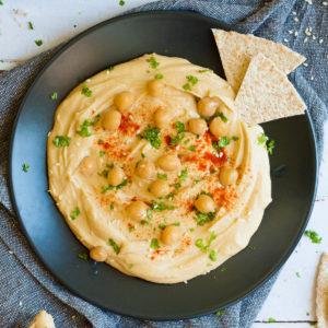 Black plate with light brown hummus sprinkled on top with chickpeas, sesame seeds, chopped parsley and paprika powder. Soft tortilla triangles are dipped at the side.