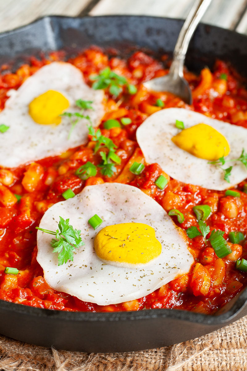 3 fried egg lookalike with white and yolk part on top of a red chunky sauce sprinkled with green spring onion rings and chopped parsley in a black cast-iron skillet