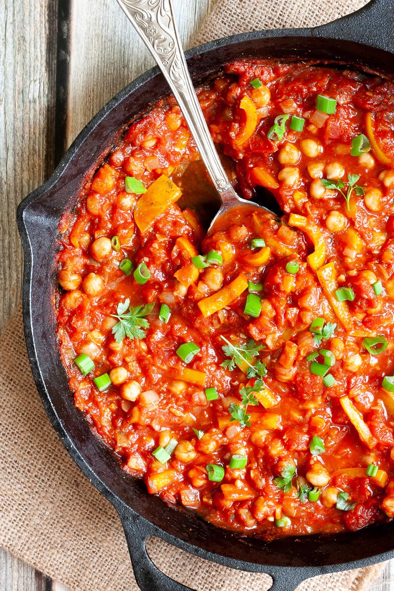 A red chunky sauce from up close sprinkled with green spring onion rings and chopped parsley. You can see chickpeas, chopped tomato and chopped yellow bell peppers.