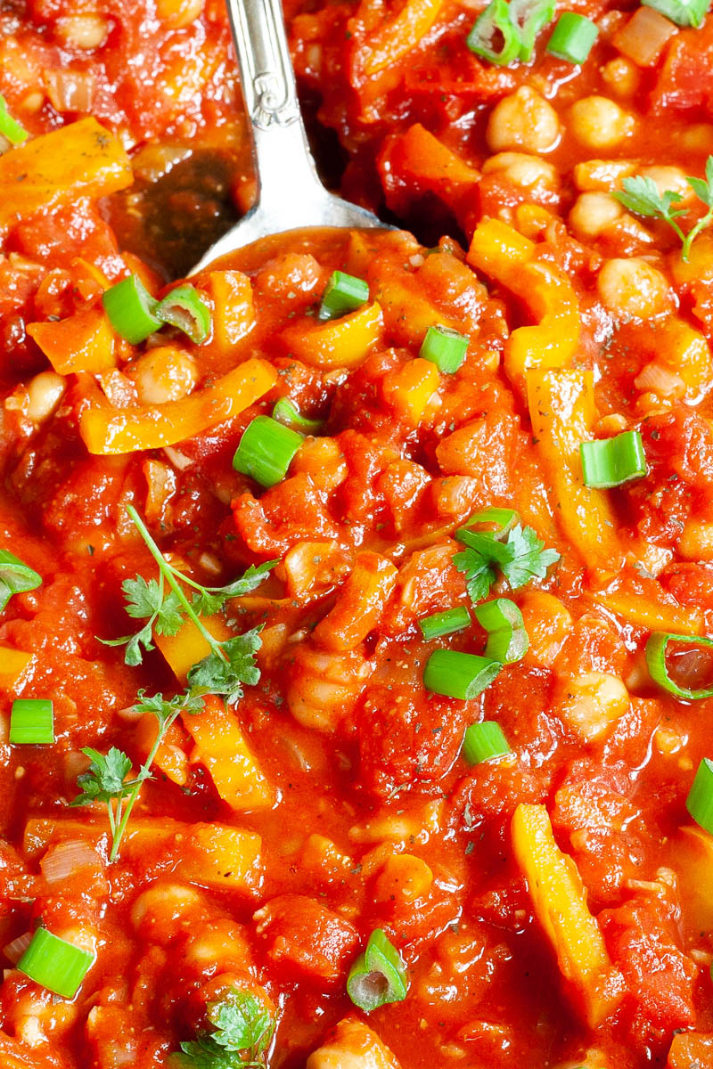 A red chunky sauce from up close sprinkled with green spring onion rings and chopped parsely. You can see chickpeas, chopped tomato and chopped yellow bell peppers.