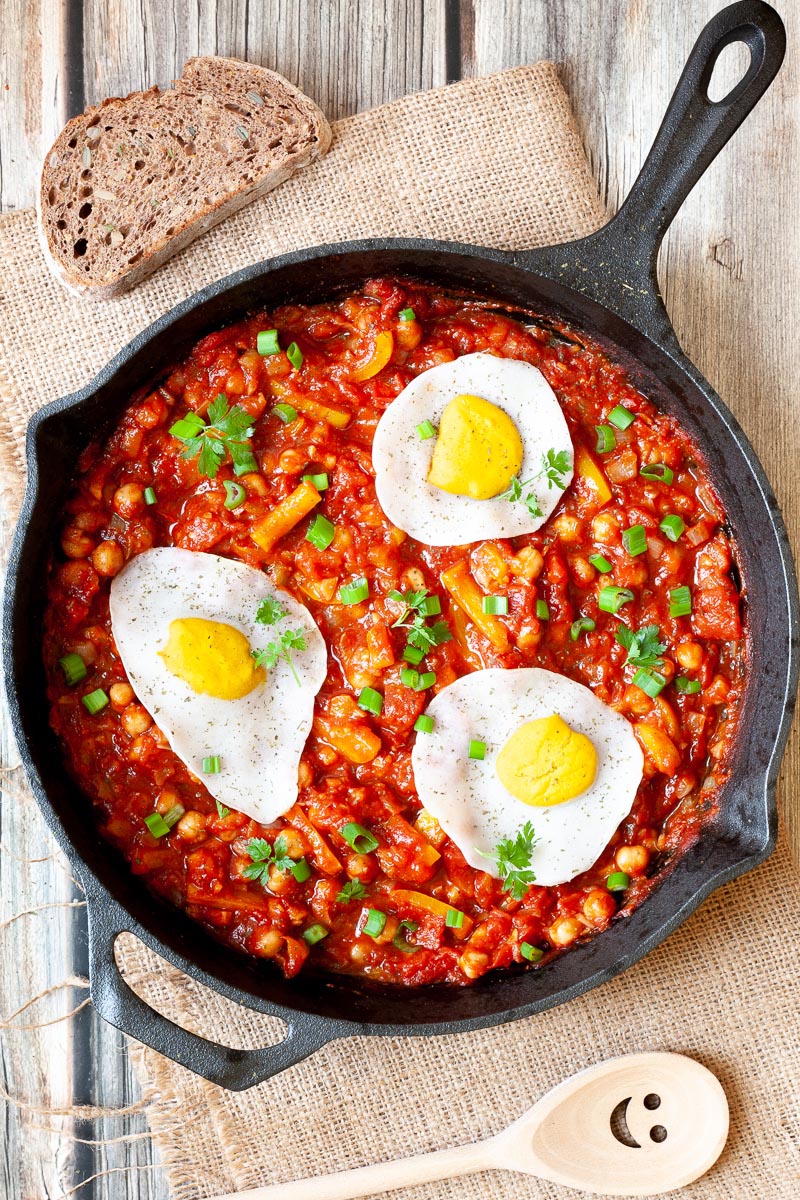 A cast iron skillet from above. 3 fried egg lookalikes with white and yolk part on top of a red chunky sauce sprinkled with green spring onion rings and chopped parsley. You can see chickpeas, chopped tomato and chopped yellow bell peppers.