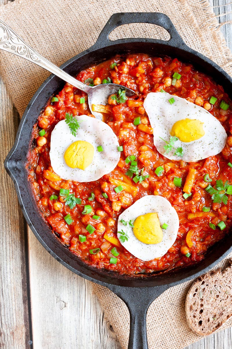 A cast iron skillet from above. 3 fried egg lookalikes with white and yolk part on top of a red chunky sauce sprinkled with green spring onion rings and chopped parsley. You can see chickpeas, chopped tomato and chopped yellow bell peppers.