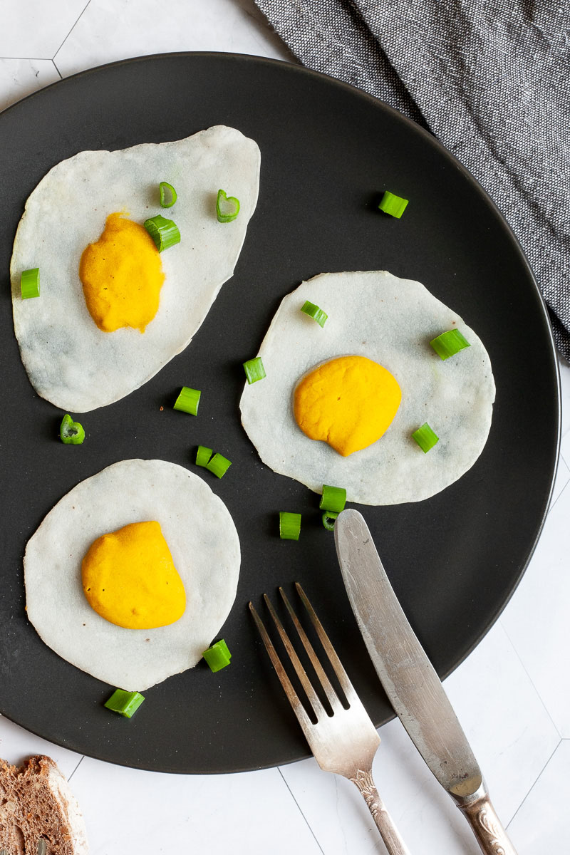 3 fried egg lookalike with white and yolk part on a black plate sprinkled with green spring onion rings.