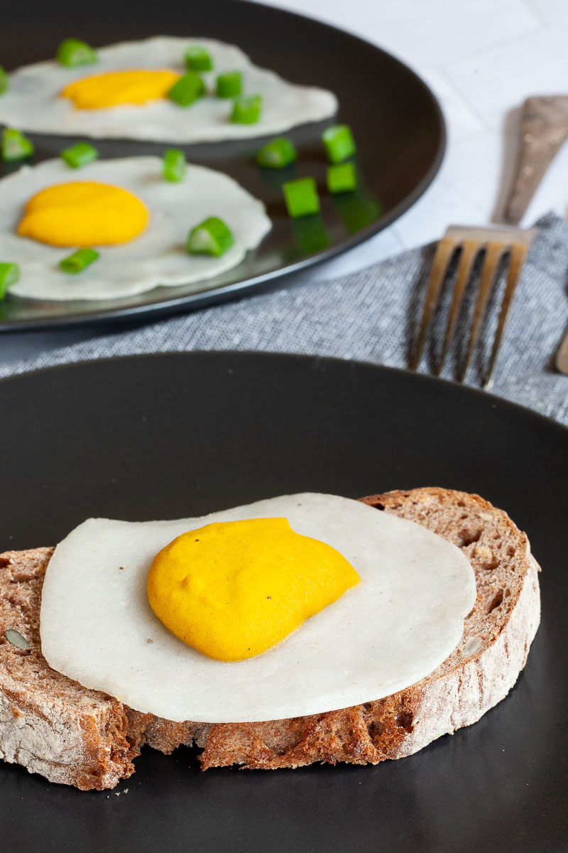 3 fried egg lookalike with white and yolk part on top a crusty brown bread served on a black plate. Fork and knife on the side. There is another black plate in the back with two more fried eggs