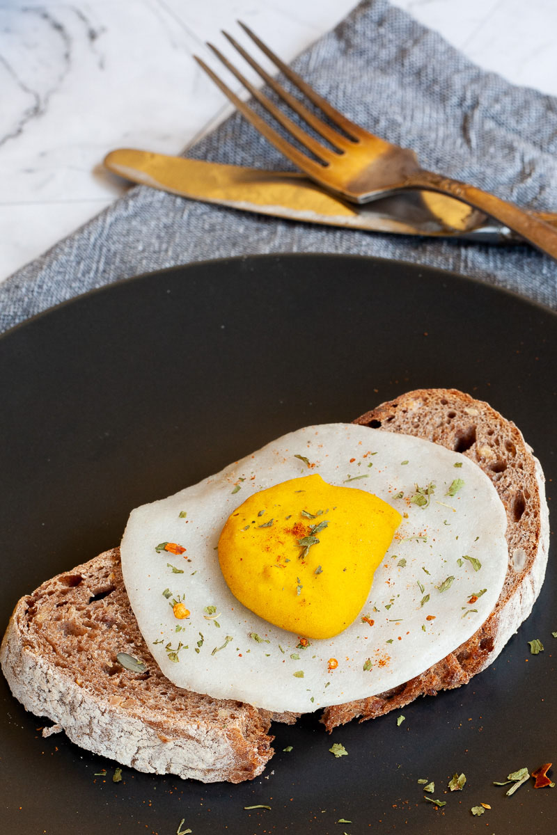 A fried egg lookalike with white and yolk part on top a crusty brown bread served on a black plate. Fork and knife on the side. It is sprinkled with dried parsley and red pepper flakes.