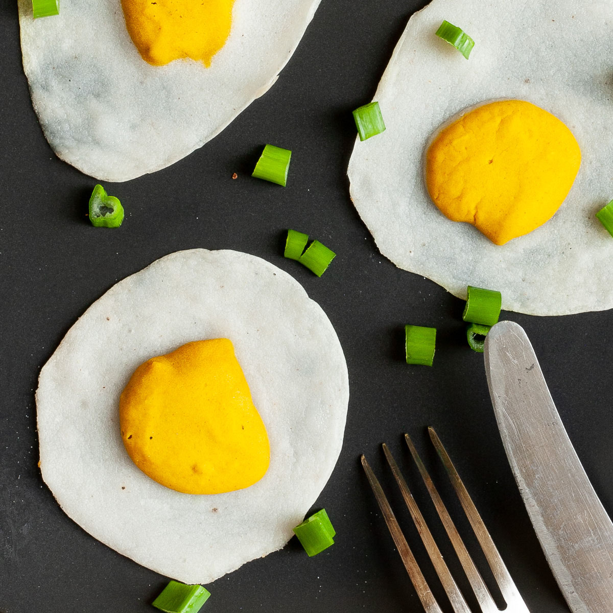 3 fried egg lookalike with white and yolk part on a black plate sprinkled with green spring onion rings.