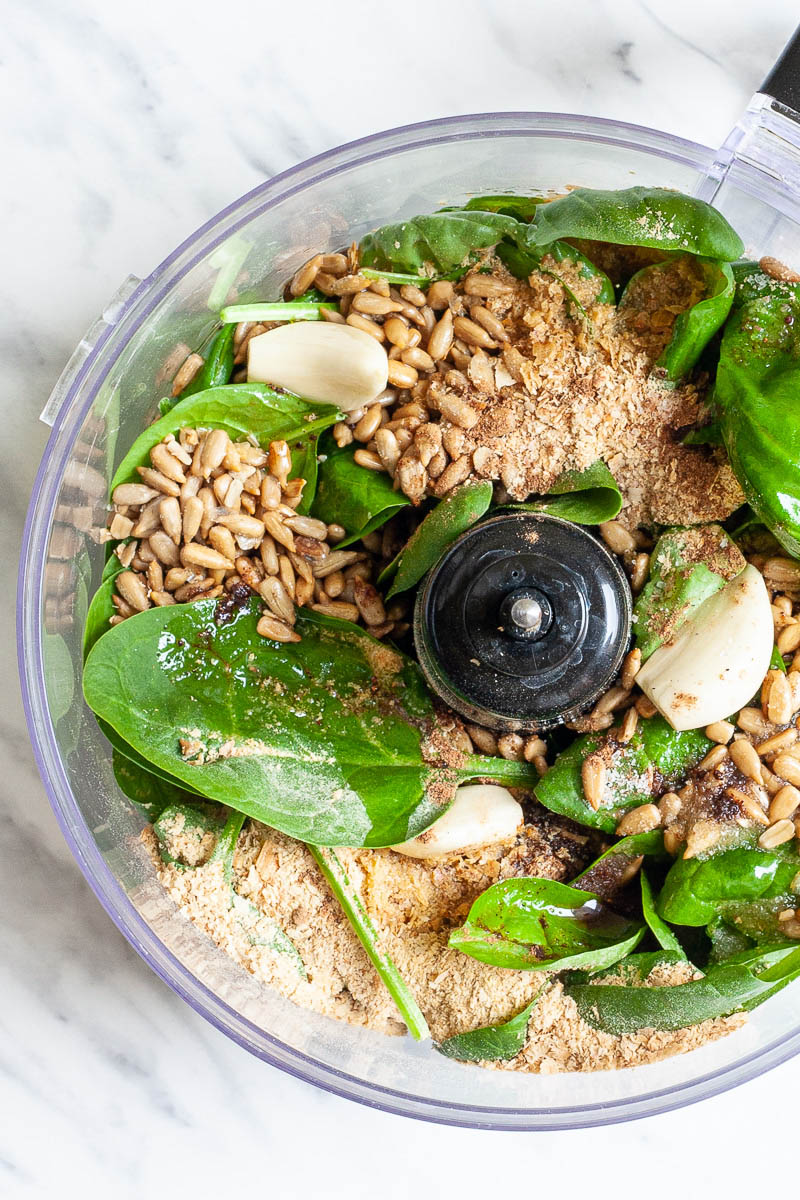 Food processor from above with sunflower seeds, fresh spinach and basil leaves, garlic clove and yellow powder to make pesto.