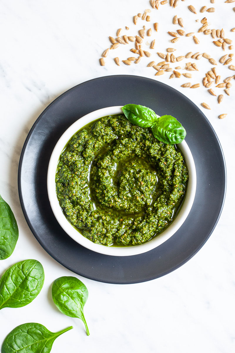 Small white bowl with a chunky green paste and a fresh basil leaf on the side. Fresh spinach leaves and sunflower seeds are scattered around it.