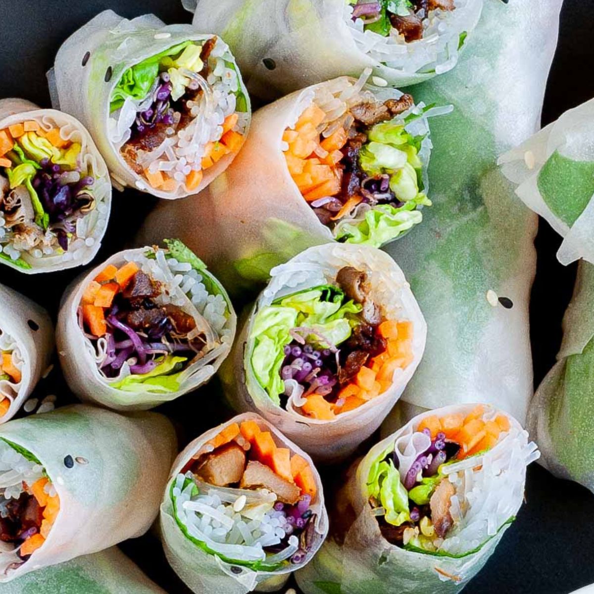 Shredded carrots, pruple cabbage and lettuce with mushroom slices and vermicelli noodles are wrapped in rice paper and cut in half. The rolls are placed on a black plate. 