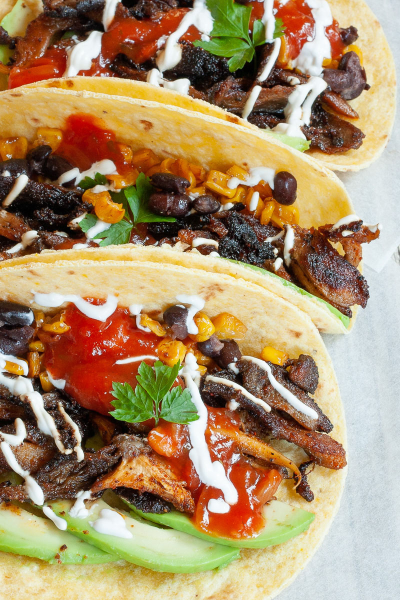 3 small tortillas folded in half  on parchment paper filled with mushroom shreds, corn, avocado slices, black beans drizzled with red and white sauce.