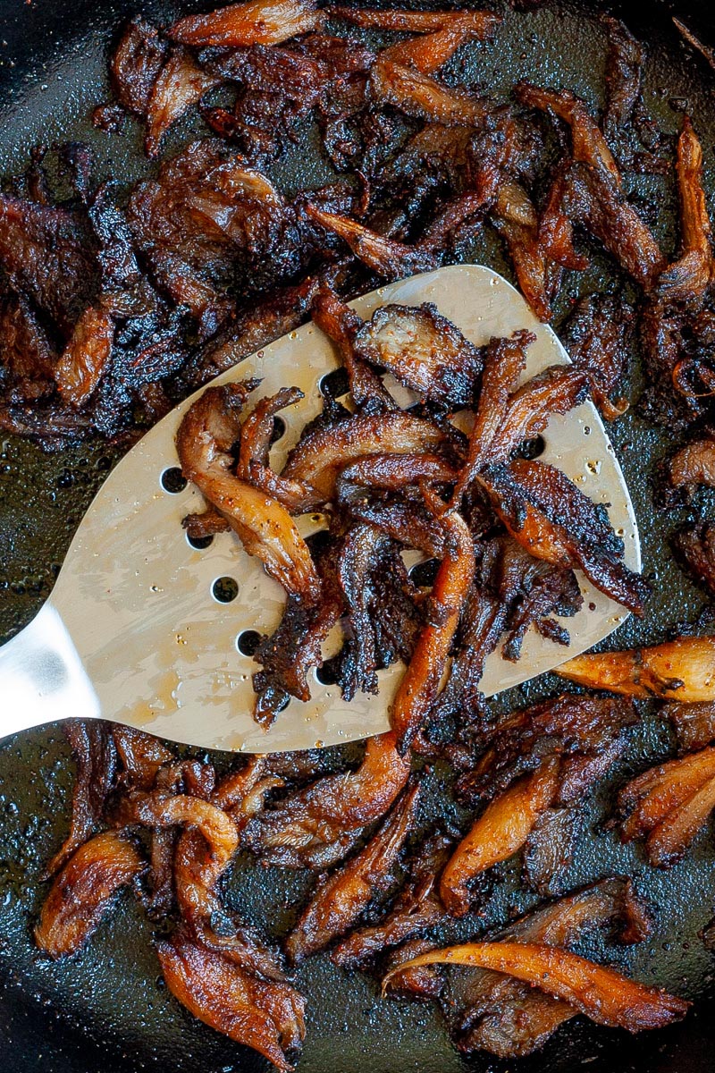 Crispy dark brown oyster mushroom shreds in a black cast-iron skillet. A spatula is holding up a handful.