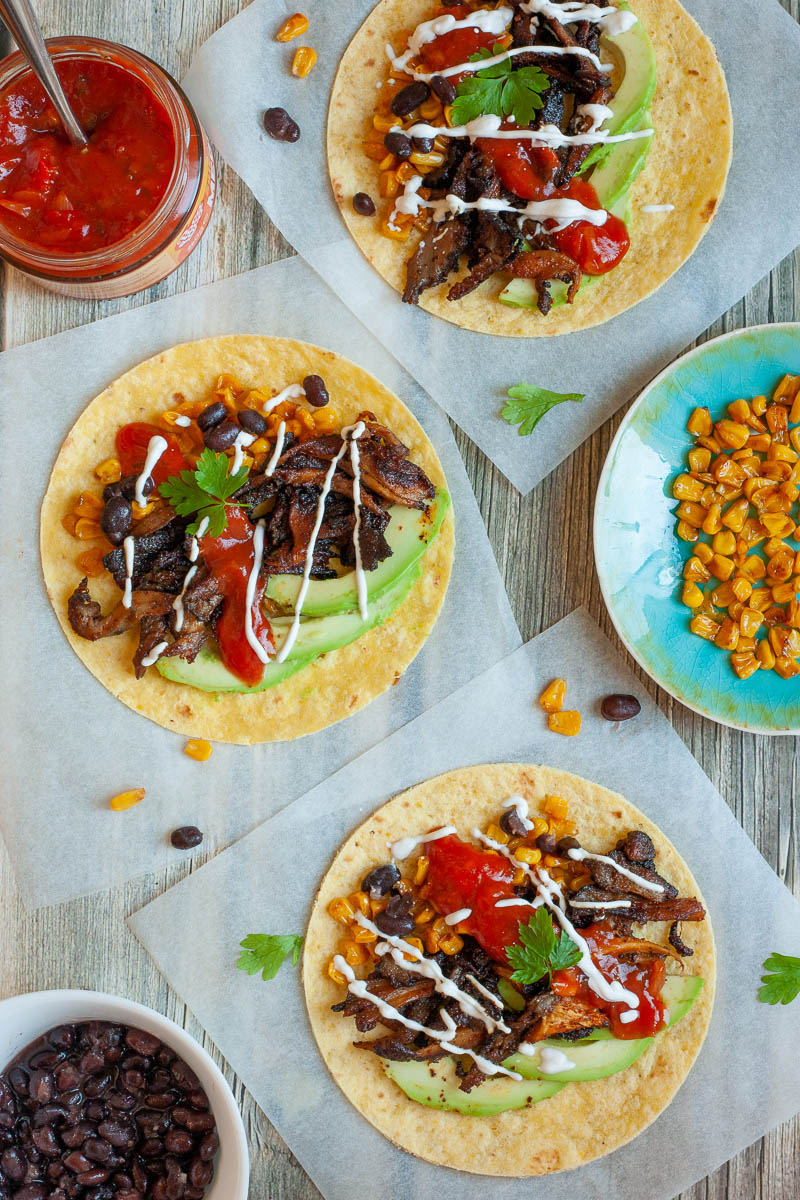 3 small tortillas on parchment paper filled with mushroom shreds, corn, avocado slices, black beans drizzled with red and white sauce. One small bowl with black beans, one with red sauce and one blue plate with corn