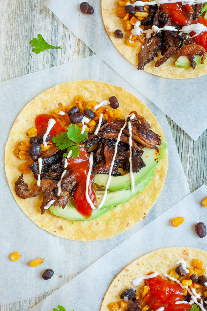 3 small tortillas on parchment paper filled with mushroom shreds, corn, avocado slices, black beans drizzled with red and white sauce.