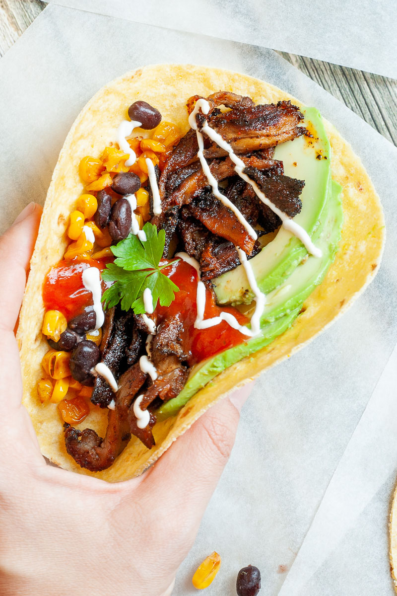 A hand is holding a small tortillas filled with mushroom shreds, corn, avocado slices, black beans drizzled with red and white sauce.