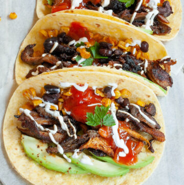 3 small tortillas folded in half on parchment paper filled with mushroom shreds, corn, avocado slices, black beans drizzled with red and white sauce.