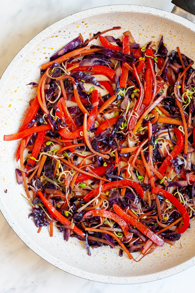 A white frying pan with shredded veggies like carrots, red cabbage, bell pepper, and bean sprouts.