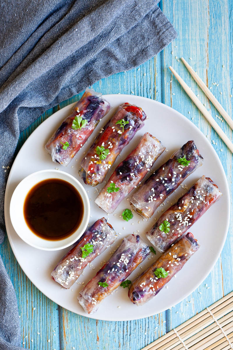 A couple of spring rolls are arranged on a white plate from above sprinkled with sesame seeds and chopped parsely. They are transparent so you can see red, orange and purple veggie pieces. A small white bowl with a dark brown dipping sauce is next to them.