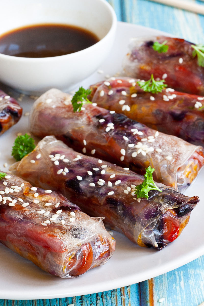 A couple of spring rolls are arranged on a white plate sprinkled with sesame seeds and chopped parsely. They are transparent so you can see red, orange and purple veggie pieces.