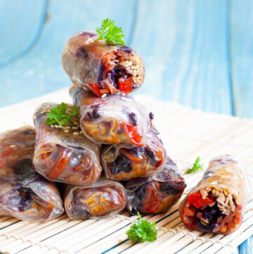 Lots of spring rolls placed on top of each other like a pyramid. The top one is cut in half so you can see inside the purple cabbage bits and some carrot and bell pepper pieces.