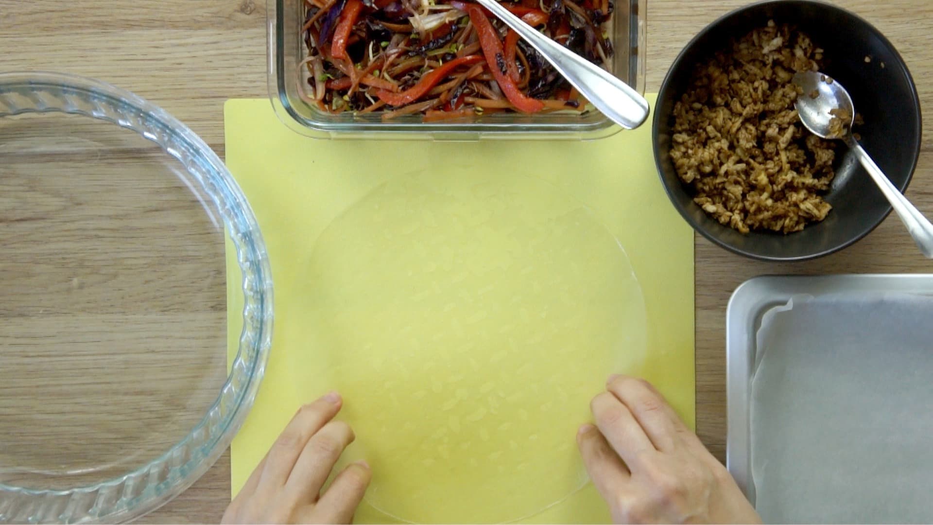 A hand places the softened rice paper on the yellow board. The fillings are in small bowls next to it.