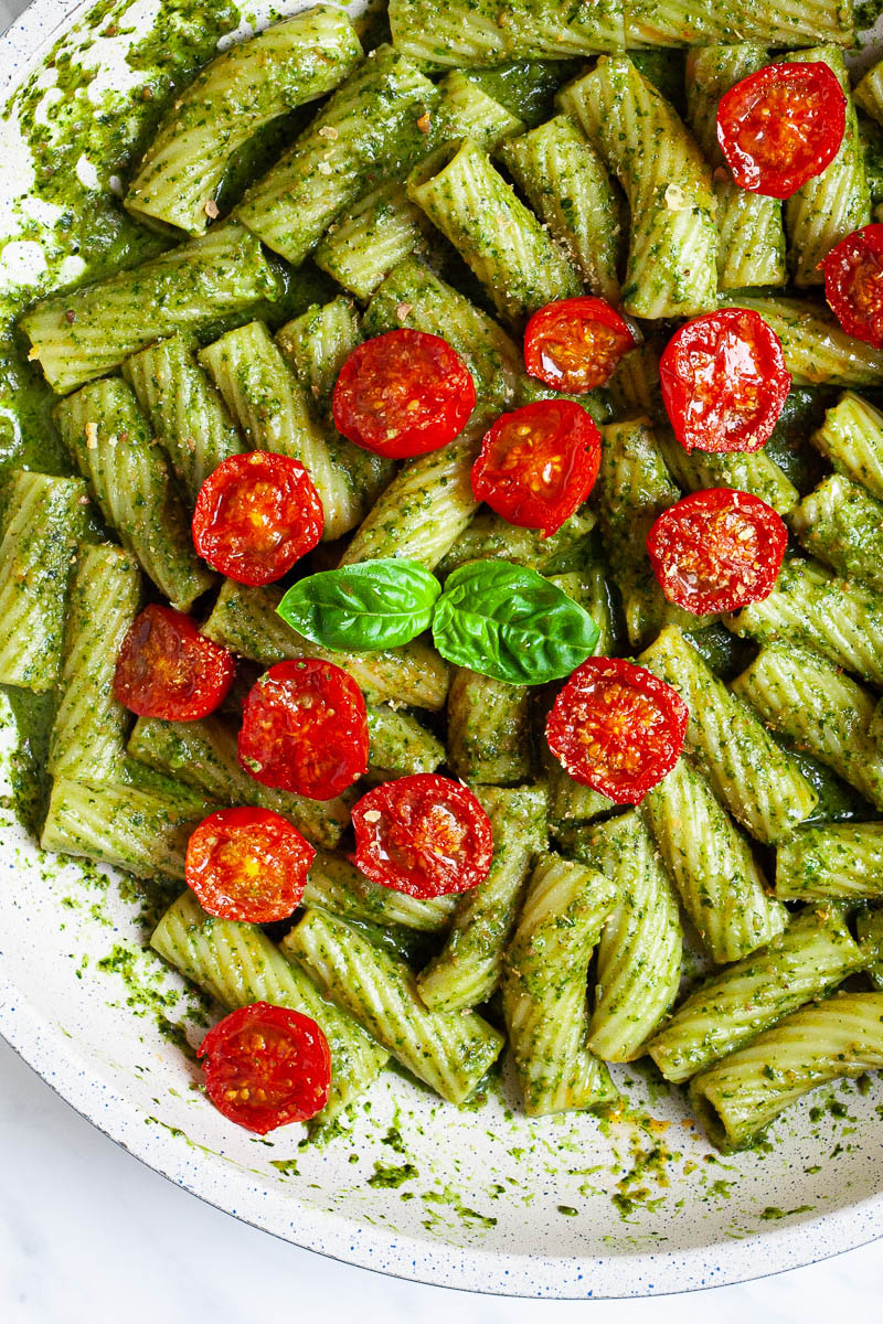 White frying pan with tortiglioni pasta covered in green paste, wilted half cherry tomatoes are on top scattered.