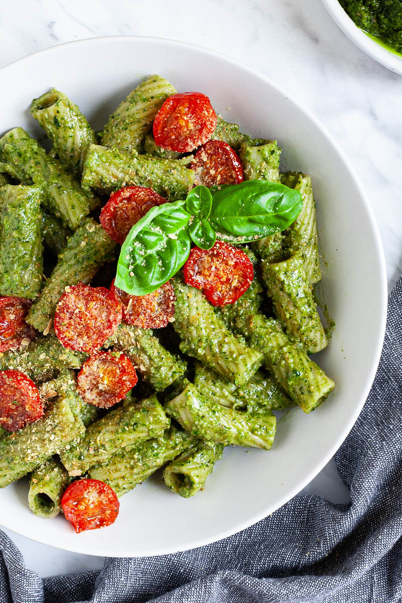 White bowl with tortiglioni pasta covered in green pesto topped with cherry tomato halves and sprinkled with yellow flakes.