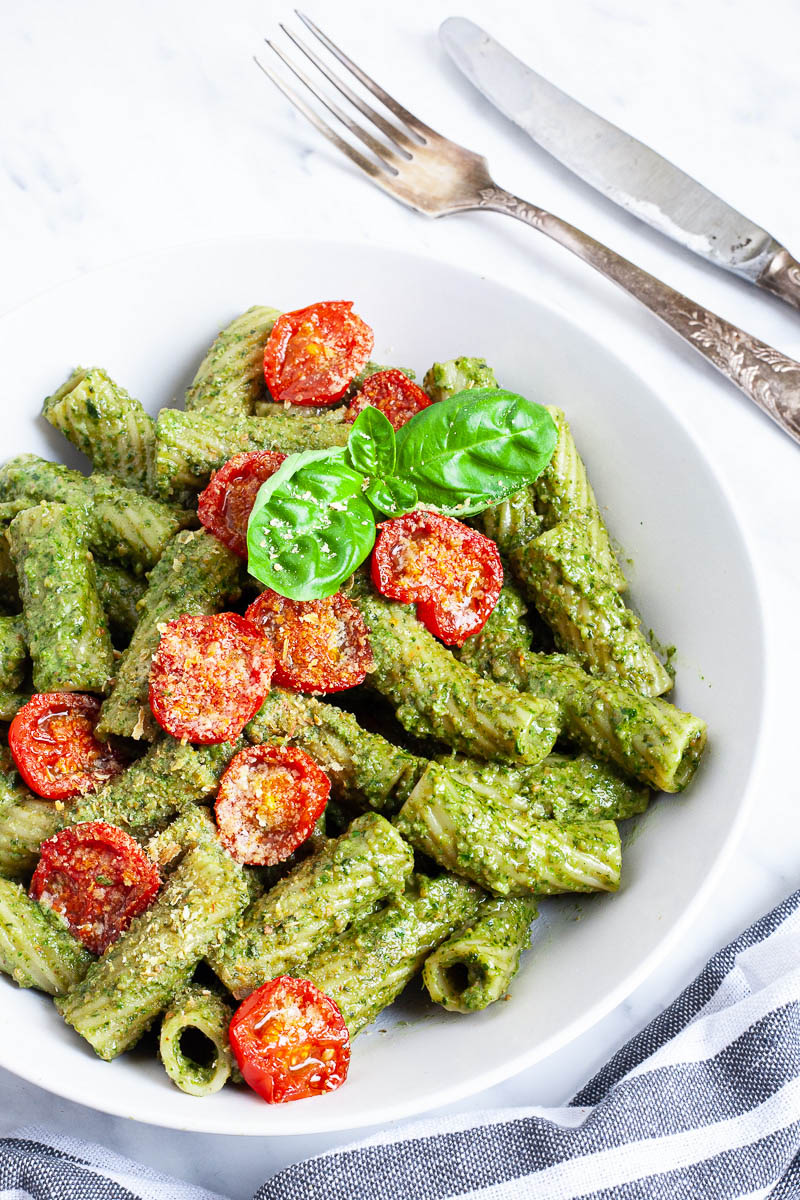 White bowl with tortiglioni pasta covered in green pesto topped with cherry tomato halves and sprinkled with yellow flakes.