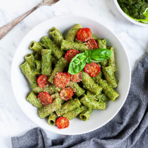 White bowl with tortiglioni pasta covered in green pesto topped with cherry tomato halves and sprinkled with yellow flakes. A small white dish is int the corner with the remaining pesto