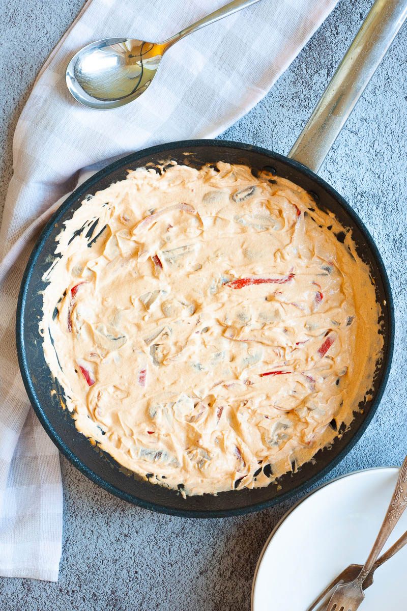 Frying pan from above with a white-orange creamy sauce with sliced crispy mushrooms, red bell pepper sticks which are almost covered in sauce.